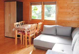 Wooden cabin interior with dining table, four chairs, gray sectional sofa and garden view through windows.