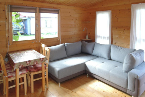 Bright living room in a wooden cabin with a gray sectional sofa, dining table, and large sunny windows in Germany.