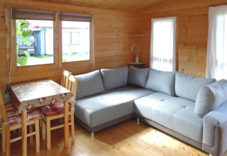 Sala luminosa en cabaña de madera en Alemania con sofá gris, mesa de comedor y grandes ventanas soleadas.