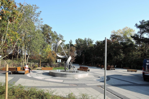 Foto de una plaza moderna con escultura y bancos, rodeada de árboles cerca de una cabaña en día soleado.