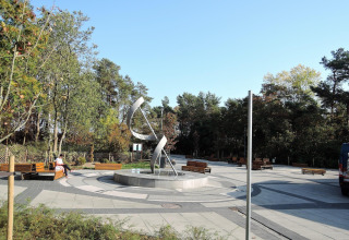 Photo of a modern plaza with sculpture and benches surrounded by trees near a cabin on a sunny day.