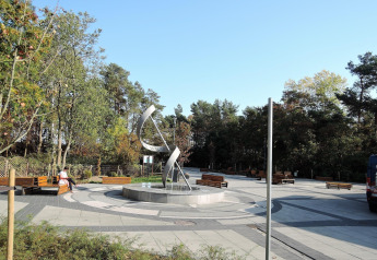 Foto de una plaza moderna con escultura y bancos, rodeada de árboles cerca de una cabaña en día soleado.
