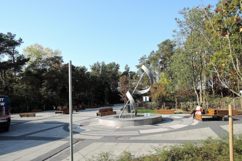 Public square with a modern metallic sculpture, benches, and trees under a clear blue sky, peaceful scene.