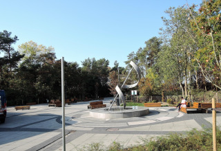 Public square with a modern metallic sculpture, benches, and trees under a clear blue sky, peaceful scene.