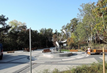 Plaza pública con escultura moderna metálica, bancos y árboles bajo un cielo azul claro, ambiente tranquilo.