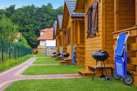 Reeks houten chalets met barbecues en blauwe plooistoelen buiten in een groene vakantieresort.