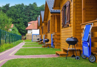 A row of wooden cabins with grills and blue folding chairs outside, set in a green holiday resort.