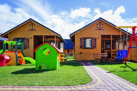 Due chalet in legno al villaggio turistico con area giochi e giocattoli colorati su prato sotto il cielo blu.
