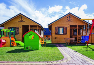Two wooden cabins at a holiday resort with playground and colorful toys on lawn under a bright blue sky.