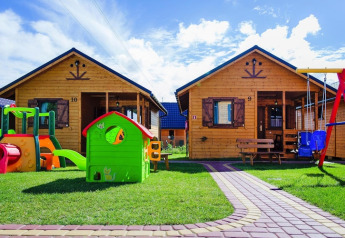 Twee houten huisjes in een vakantieresort met speeltuin en kleurrijke speeltoestellen op het gras onder blauwe lucht.
