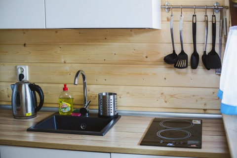 Modern wooden kitchen with electric kettle, dish soap, black sink, cooktop, and hanging utensils set.