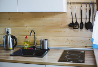 Modern wooden kitchen with electric kettle, dish soap, black sink, cooktop, and hanging utensils set.