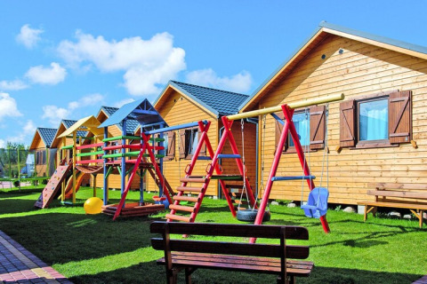 Holiday resort with wooden lodges, colorful playground, swings and benches on green grass in sunny weather.