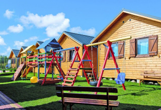Holiday resort with wooden lodges, colorful playground, swings and benches on green grass in sunny weather.