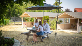 Drei Personen sitzen an einem Picknicktisch unter einem Sonnenschirm vor Ferienhäusern in Miedzyzdroje.