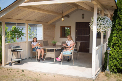 Family sits on covered terrace at holiday home in Miedzyzdroje, Poland, surrounded by greenery and flowers.