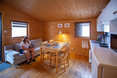 Interieur van een knusse houten blokhut met bank, eettafel, kleine keuken en een vrouw op de bank.