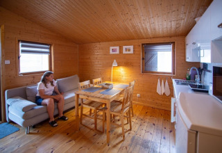 Interieur van een gezellige houten chalet met zetel, eettafel, compacte keuken en vrouw op de zetel.
