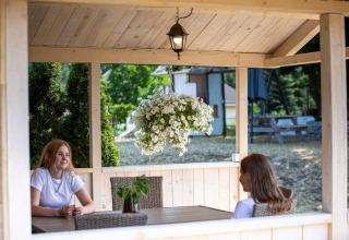 Two women sit in a wooden cabin with hanging flowers, surrounded by nature at Holiday homes in Miedzyzdroje, Germany.