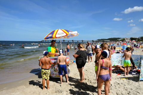 Kinderen en volwassenen verzamelen zich bij een ijsstand op een druk strand met kleurrijke windschermen.