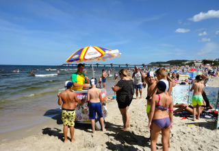 Kinderen en volwassenen verzamelen zich bij een ijsstand op een druk strand met kleurrijke windschermen.
