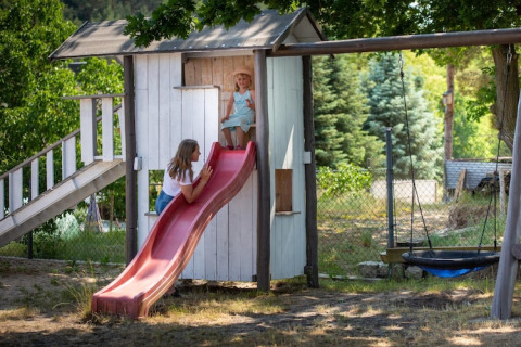 Two children play on a red slide attached to a small wooden cabin with a swing, surrounded by trees.