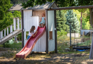 Twee kinderen spelen op een rode glijbaan van een houten speelhuisje met schommel en veel groen.
