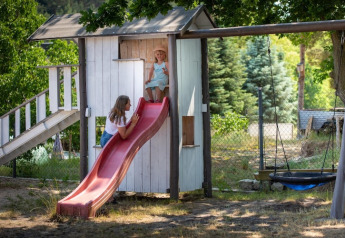 Zwei Kinder spielen an einer Rutsche bei einem Baumhaus mit Schaukeln, umgeben von Bäumen und Gartenzaun.