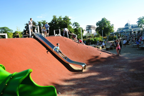 Kinderen spelen op een speelplein met glijbaan en klimhelling bij een vakantiehuisje op een zonnige dag.
