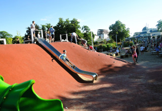 Kinderen spelen op een speelplein met glijbaan en klimhelling bij een vakantiehuisje op een zonnige dag.
