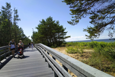 Houten wandelpad aan het strand met mensen, kinderwagen en dennen nabij vakantiehuizen in Miedzyzdroje.