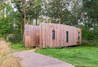 Modern wooden glamping cabin surrounded by lush greenery and trees, viewed from the pathway entrance.