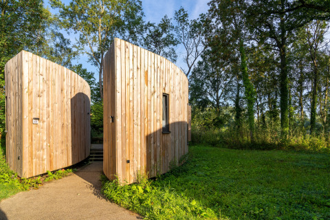 Wood-clad tiny house named The Lodge by YourNature, surrounded by greenery and forest in sunlight.
