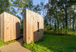 Træbeklædt tiny house kaldet The Lodge af YourNature, omgivet af grønt og skov i sollys.