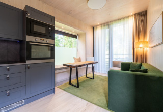 Modern glamping accommodation with green sofa, kitchen area, dining table, large windows, and cozy lighting.