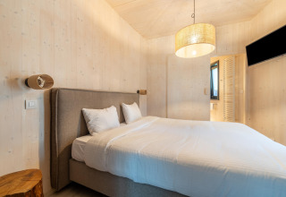 Moderne glamping-indkvartering med dobbeltseng, trævægge, natbord og vægmonteret tv i soveværelset.