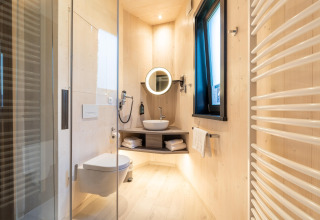 Moderne badeværelse i glamping-indkvartering med lyse trævægge, bruser, væghængt toilet og vindue.