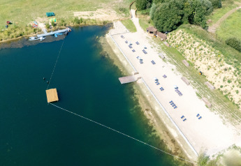 Vue aérienne d’un site de glamping avec plage de sable, transats et kayaks au bord d’un lac paisible.