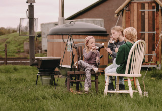 Three children roast marshmallows by a fire outside a wooden hut at Feather Down De Oude Melkstal holiday park.