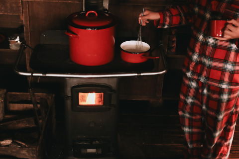 Personne en pyjama à carreaux rouges cuisinant sur poêle à bois avec casseroles rouges dans un chalet.