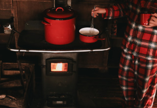 Person in red plaid pajamas cooking on a wood stove with red pots in a cozy wooden cabin interior.