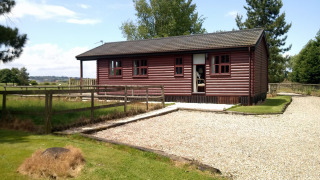 Foto del chalet Five Waterings, cabaña de madera con jardín vallado, camino de grava y entorno rural.