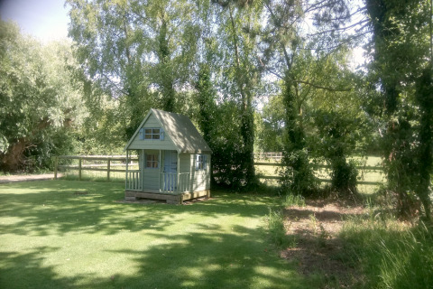 Lille grønt legehus ved 'Five Waterings' chalet i England, omgivet af træer og frodig græsplæne.