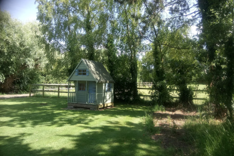 Lille grønt legehus ved 'Five Waterings' chalet i England, omgivet af træer og frodig græsplæne.