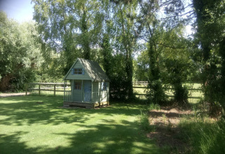 Lille grønt legehus ved 'Five Waterings' chalet i England, omgivet af træer og frodig græsplæne.