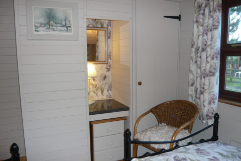 Soveværelse i Five Waterings chalet med fletstol, spejl, skammel og vindue med blomstrede gardiner.