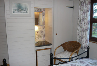Soveværelse i Five Waterings chalet med fletstol, spejl, skammel og vindue med blomstrede gardiner.
