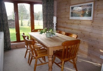 Comedor con mesa de madera, seis sillas y grandes ventanales con vista al jardín en Rainbow Petty Lodge UK.