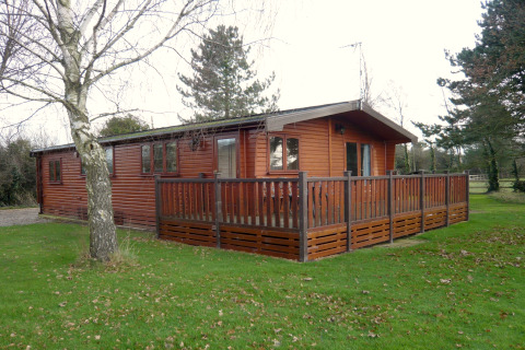 Cabaña Rainbow Petty en Reino Unido, construida en madera, con terraza vallada y rodeada de árboles y césped.