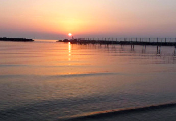 Zonsondergang boven kalm water gezien vanuit een stacaravan, met een lange pier en zachte kleuren.